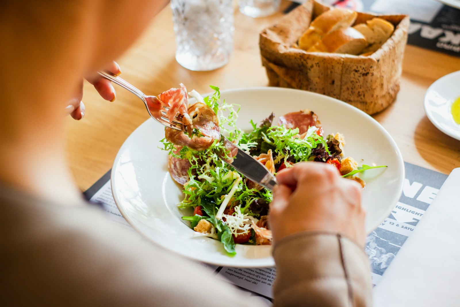 Close view of hands using a fork and knife with a salad on a table beside bread.