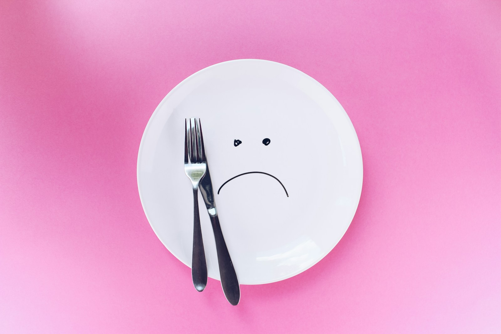 A white plate with a sad face beside cutlery on a pink surface.