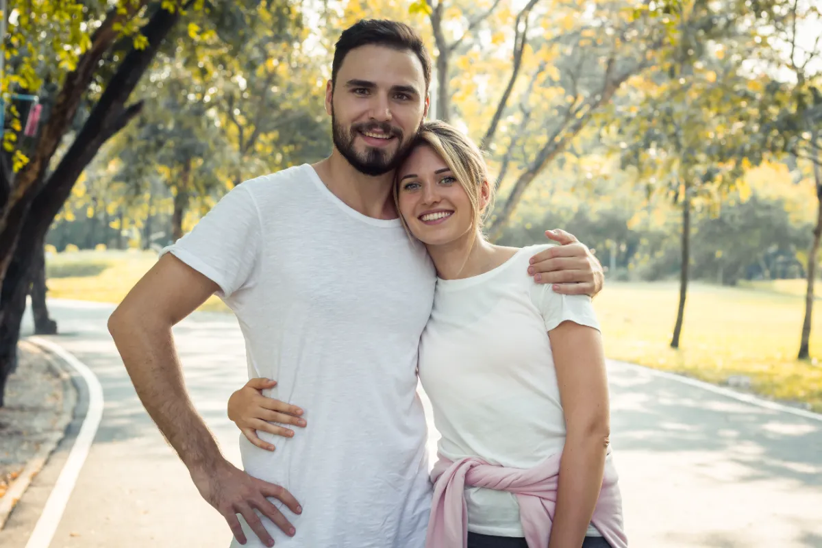 Fully clothed adult couple stands together in a leafy park.
