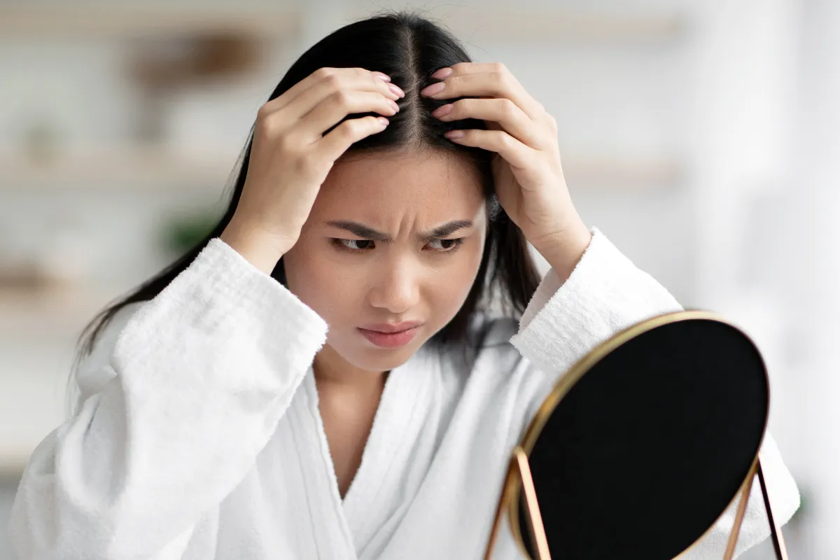 Adult woman parts her hair while looking at her scalp in a mirror.