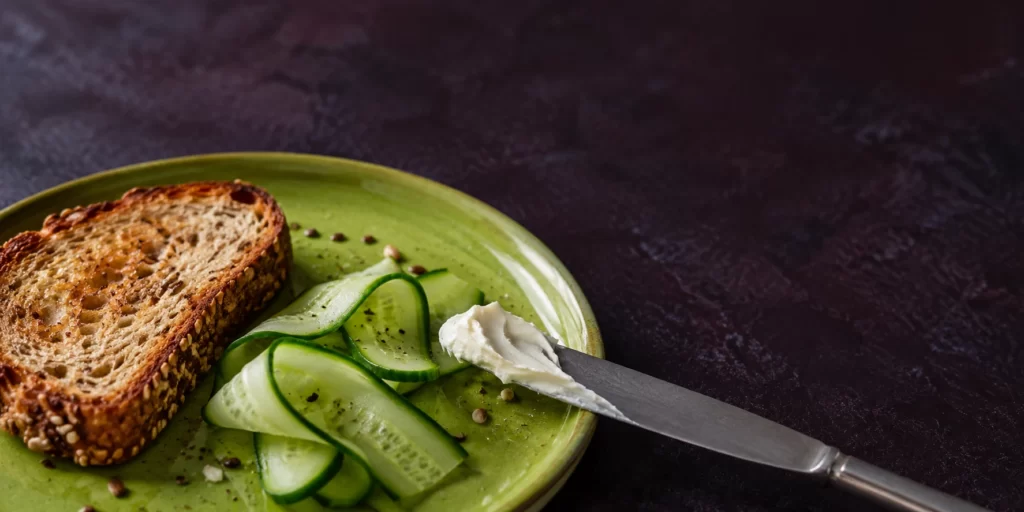 Original AI-generated editorial image: Cream-cheese smear on a knife beside seeded toast and cucumber on a lime-green plate.