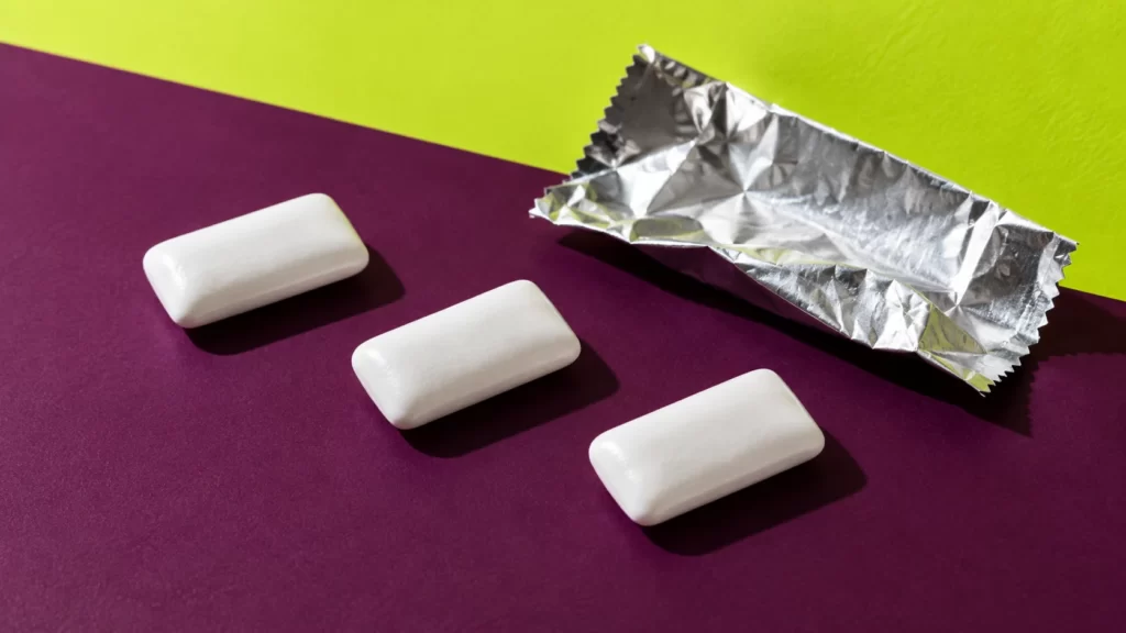 Three white rectangular chewing-gum pieces and an unbranded foil wrapper on plum and citrus-lime studio surfaces.