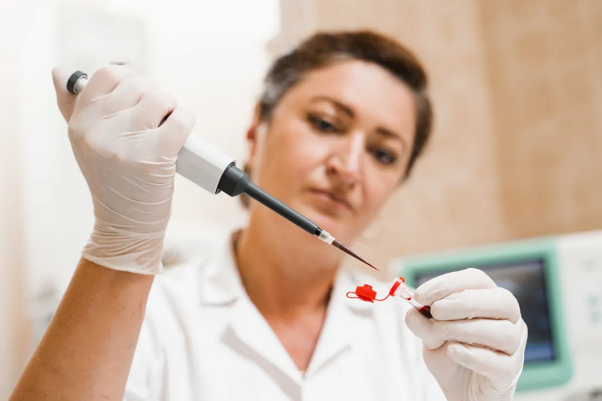 Gloved laboratory professional uses a pipette above a blood-sample tube.