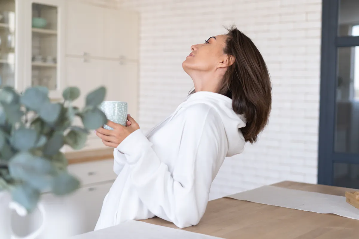 An adult in a white top holding a mug beside a kitchen counter.