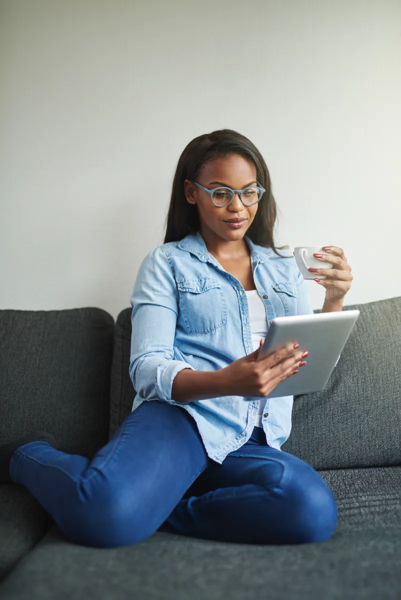 An adult using a tablet while holding a cup on a sofa.