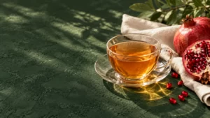 Amber tea in a glass cup beside a pomegranate on a deep-green surface with ivory linen.