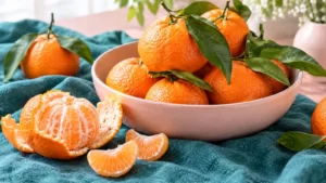 Fresh mandarins, green leaves and peeled segments beside a pink bowl on teal linen.
