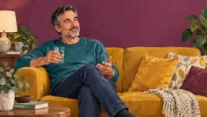 Gouache illustration of a man in a teal sweater holding water on a mustard sofa against a plum wall.