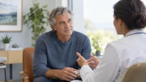 A fictional older man in a slate-blue sweater speaks with a clinician in a consultation room.